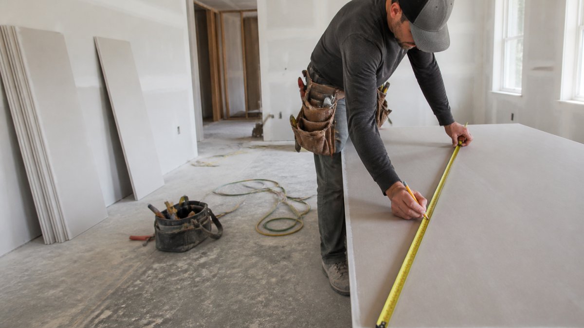 Professional contractor measuring and marking drywall for installation in a residential interior, with natural light from windows illuminating the workspace, tools and materials visible on the floor